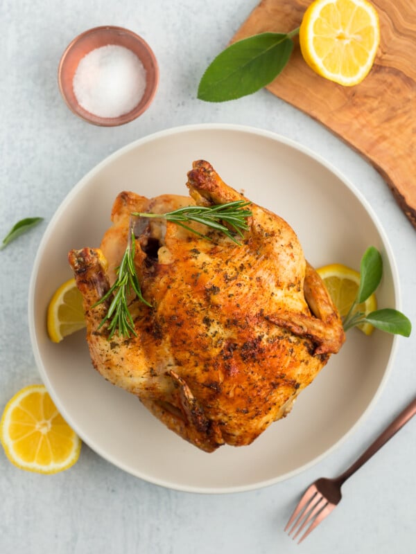 a roasted air fryer cornish hen on a white plate with sage, lemon, and a bowl of salt.