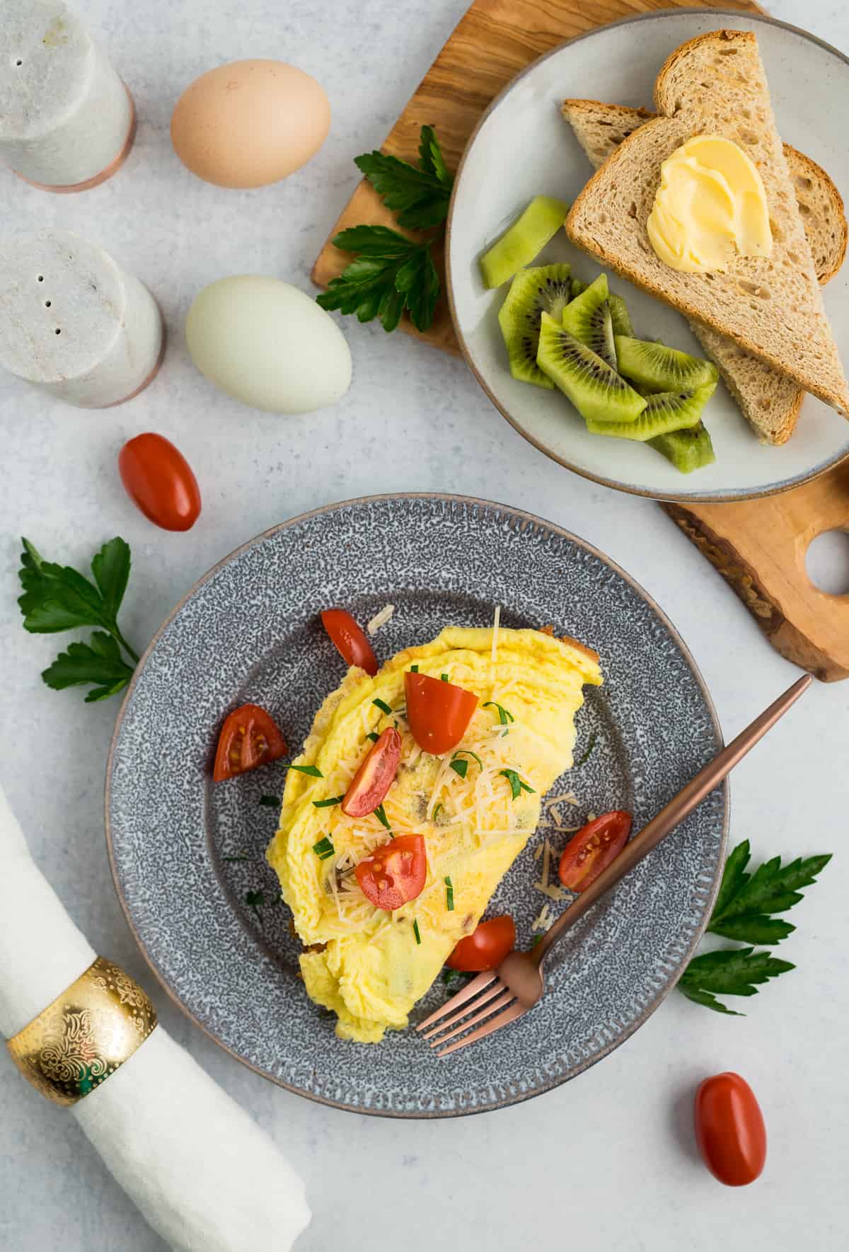 omelette on a grey plate topped with tomatoes and another plate with toast and kiwi