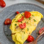 a fluffy air fryer omelet topped with sliced tomatoes, cheese, and parsley on a grey plate.