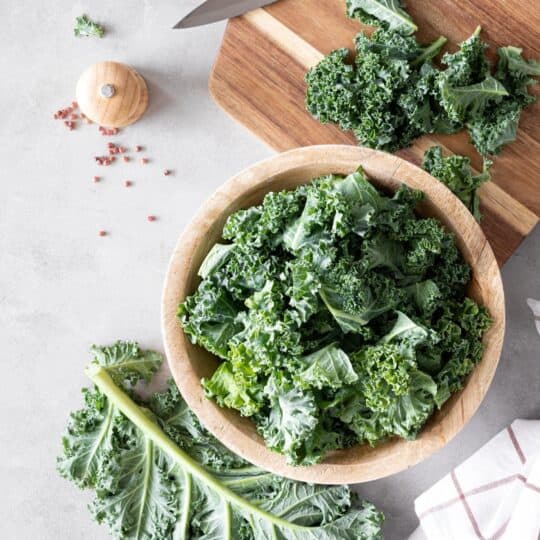 a wooden bowl full of chopped kale, and a wooden cutting board with a knife.