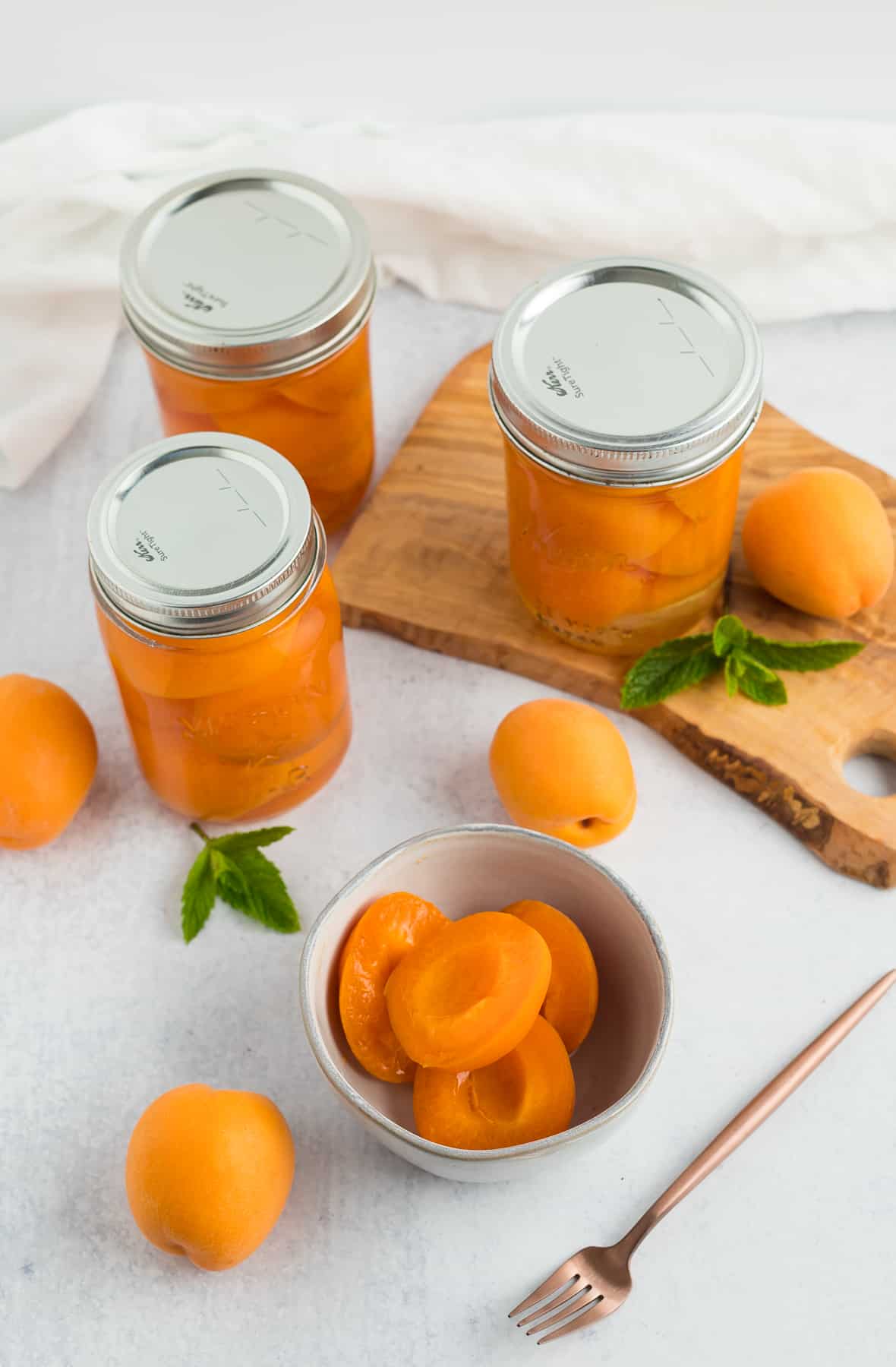 3 glass jars of preserved apricots with a few sliced apricots in a grey bowl.