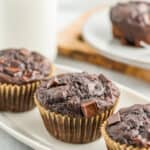 3 chocolate zucchini muffins on a white plate with a small jug of milk behind it.