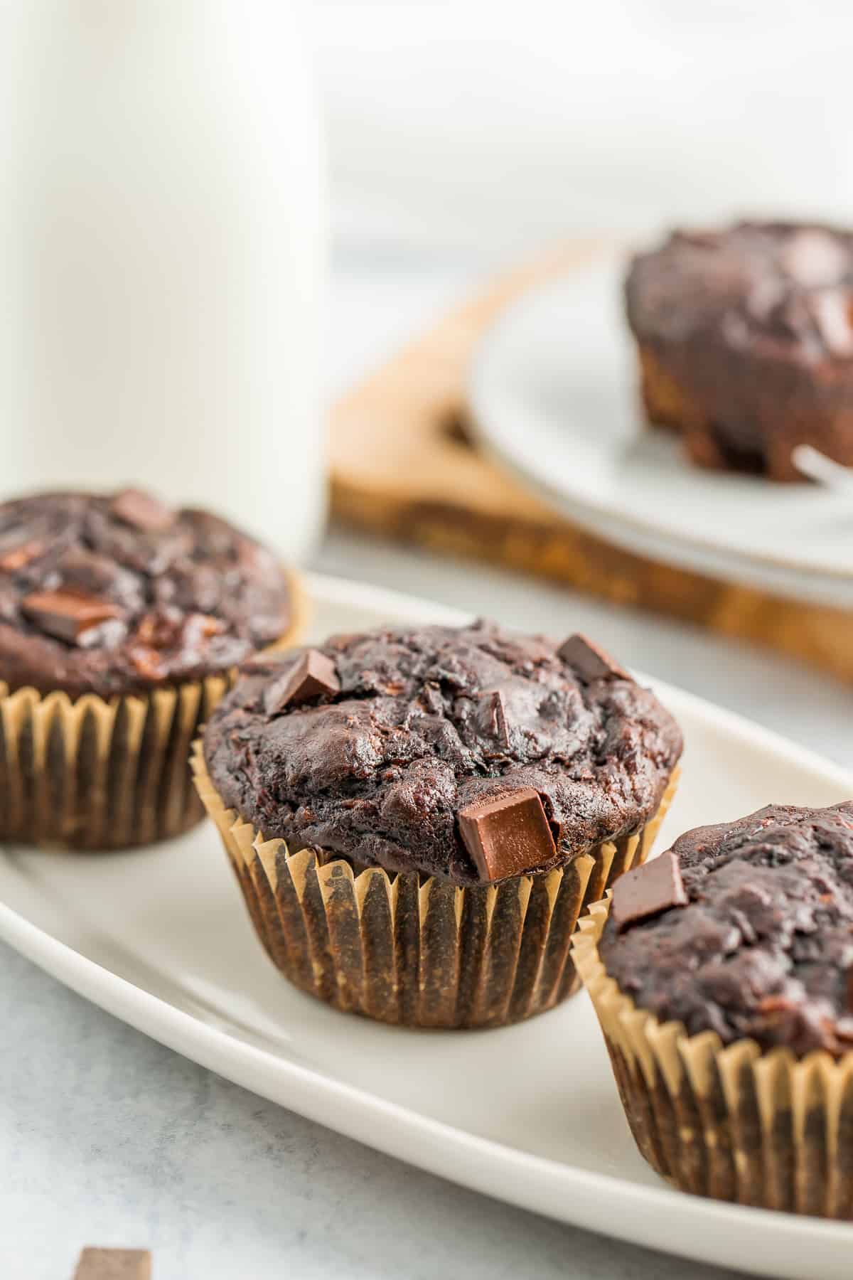 3 chocolate zucchini muffins on a white plate with a small jug of milk behind it.