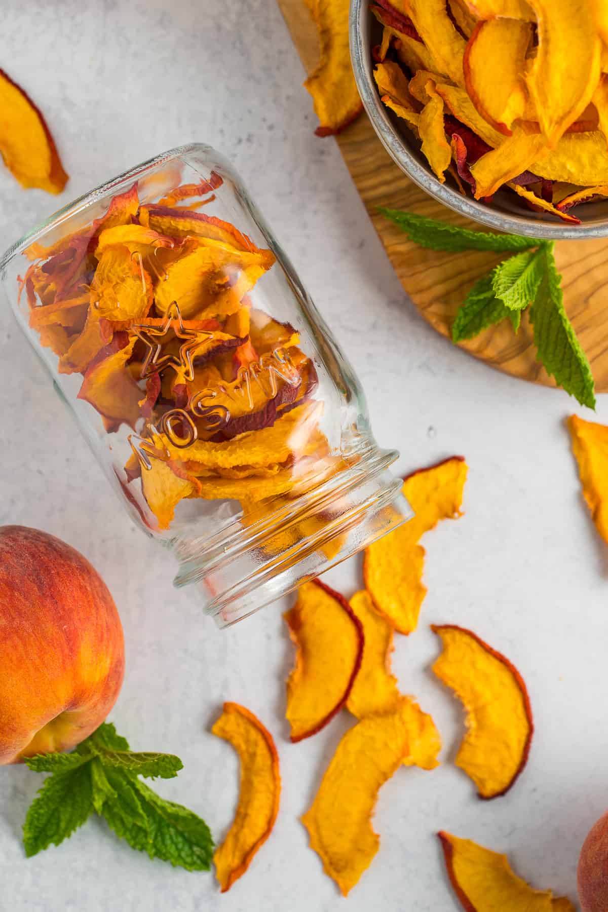a mason jar of dehydrated peach slices on a grey board with fresh peaches and mint.