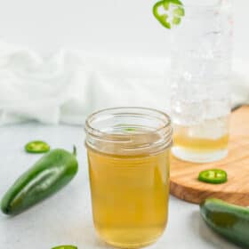 a half-pint canning jar of jalapeño simple syrup with a glass of sparkling water and jalapeños on a white board.