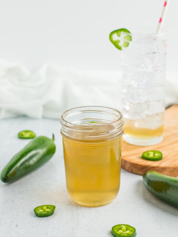 a half-pint canning jar of jalapeño simple syrup with a glass of sparkling water and jalapeños on a white board.