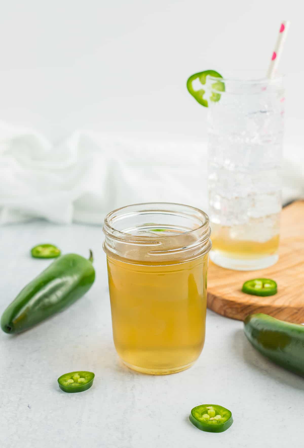 a half-pint canning jar of jalapeño simple syrup with a glass of sparkling water and jalapeños on a white board.