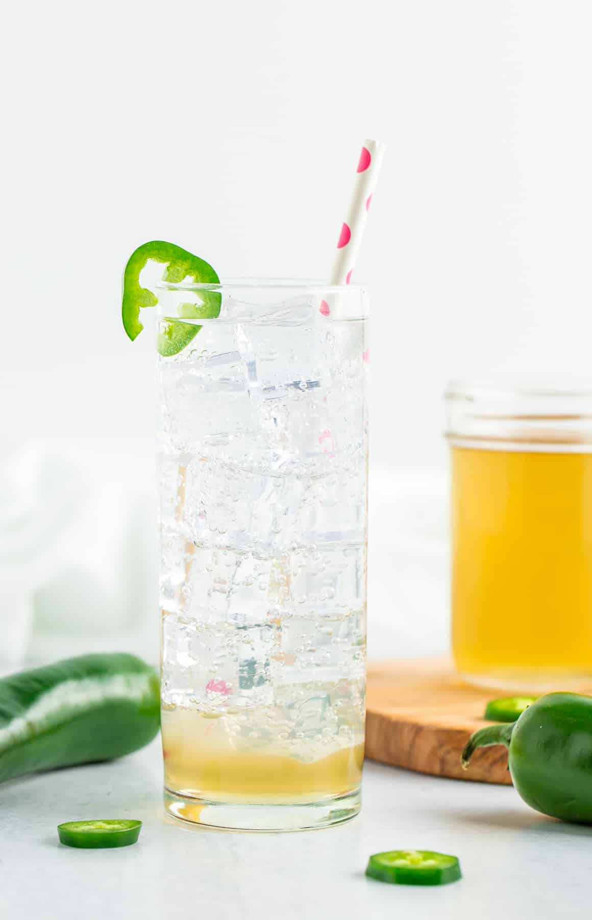 a tall glass of sparkling water with a straw and a slice of jalapeño on a white board.