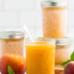 4 jars of peach freezer jam with fresh peaches and mint on a white board.