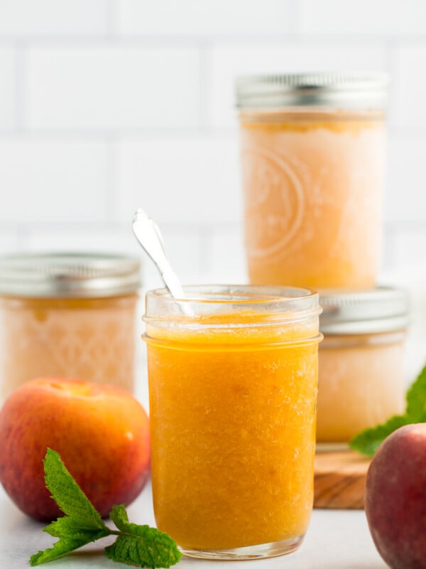 4 jars of peach freezer jam with fresh peaches and mint on a white board.