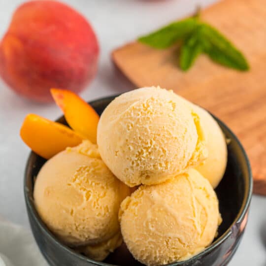 a blue bowl with multiple scoops of homemade peach ice cream, with whole peaches.