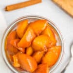 a glass jar of peach compote on a white board with a spoon and cinnamon sticks.