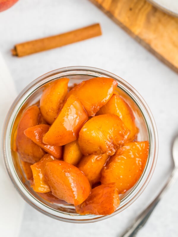 a glass jar of peach compote on a white board with a spoon and cinnamon sticks.