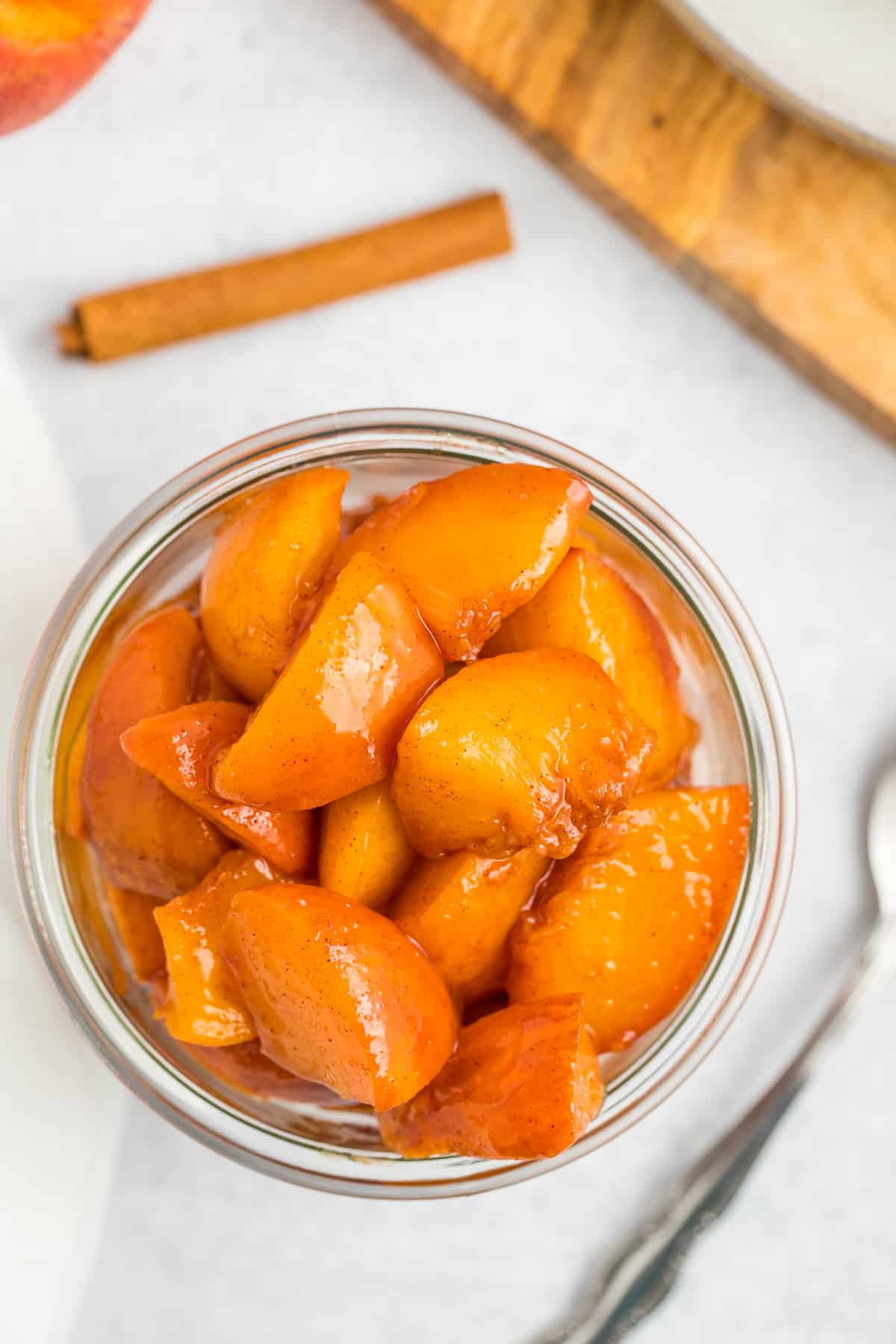 a glass jar of peach compote on a white board with a spoon and cinnamon sticks.