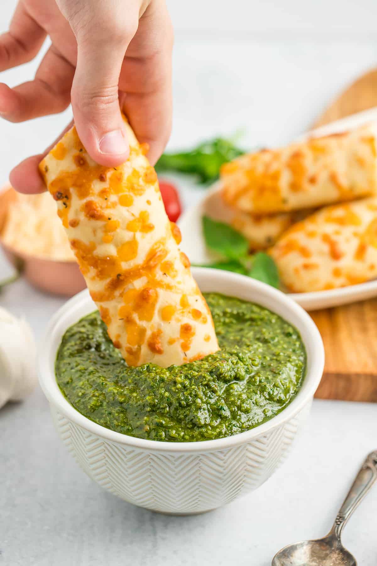 a hand dipping a cheesy breadstick into a white bowl of homemade pesto.