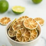 a white bowl of dried lime slices on a marble board with whole and halved limes.