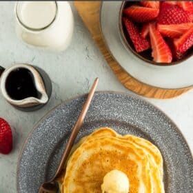 a grey plate with pancakes, a fork, and butter on a grey board with a bowl of strawberries and small container of milk.