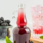 a glass bottle of blackberry simple syrup with a glass of syrup and sparkling water with a gold straw.