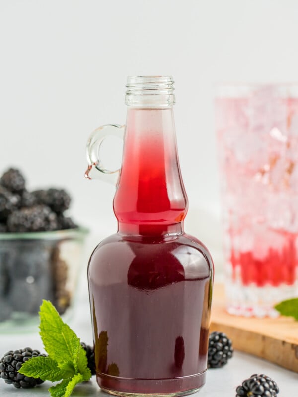 a glass bottle of blackberry simple syrup with a glass of syrup and sparkling water with a gold straw.