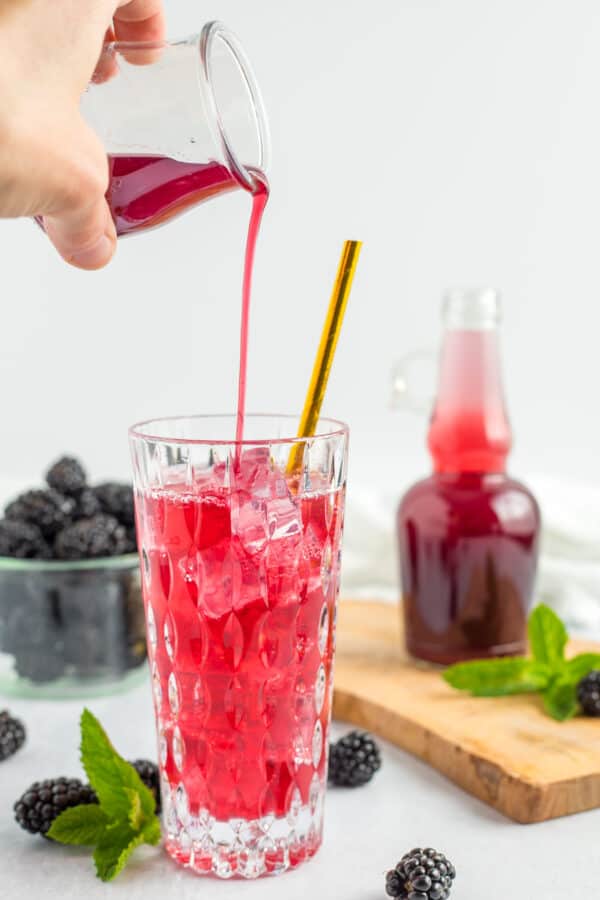 a hand pouring blackberry syrup into a glass with a gold straw.