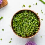 a copper bowl of dried chopped chives on a white board with a purple chive blossom.