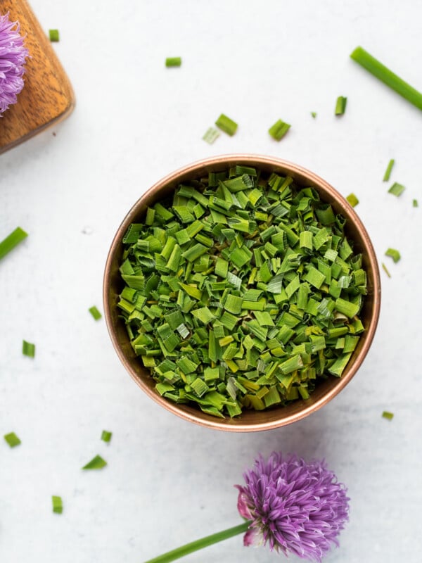 a copper bowl of dried chopped chives on a white board with a purple chive blossom.