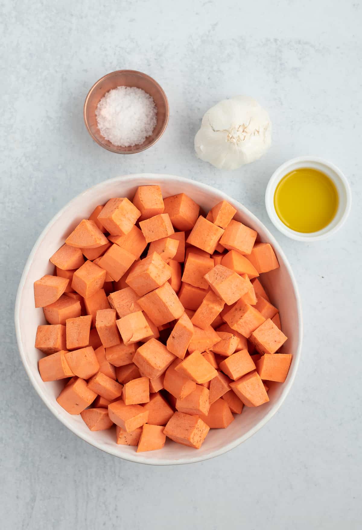 raw sweet potato cubes in a bowl with a bowl of salt, garlic, and a bowl of olive oil.