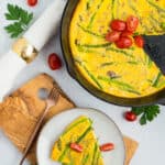 a slice of dairy-free frittata on a grey plate with the rest in a cast iron skillet topped with sliced tomatoes.