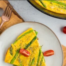 a slice of dairy-free frittata on a grey plate with the rest in a cast iron skillet topped with sliced tomatoes.