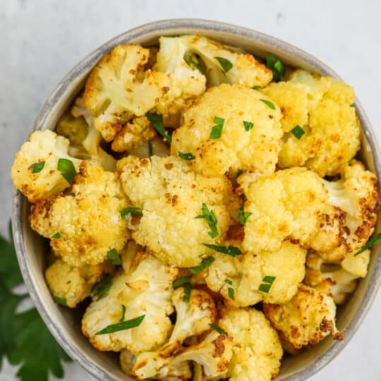 a grey bowl of air fryer cauliflower topped with chopped parsley on a grey board.