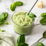 a glass bowl filled with mayonnaise mixed with pesto, with basil and cheese on the white board.
