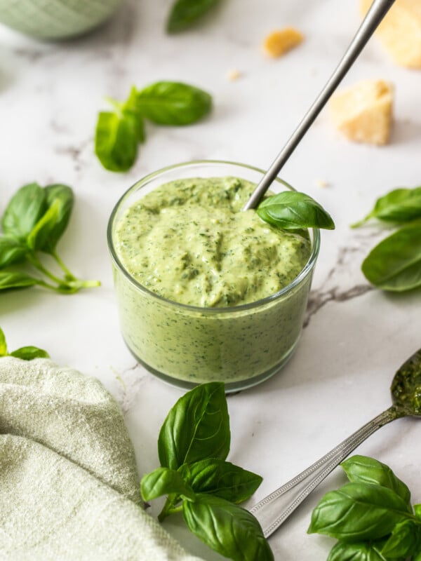 a glass bowl filled with mayonnaise mixed with pesto, with basil and cheese on the white board.