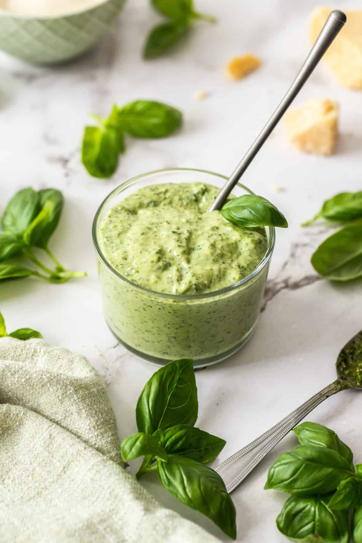 a glass bowl filled with mayonnaise mixed with pesto, with basil and cheese on the white board.