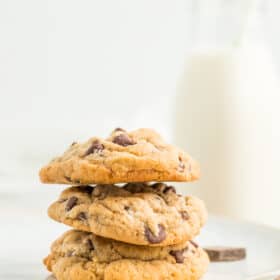 3 chocolate chip cookies stacked on a plate with a small bottle of milk with a straw in the background.