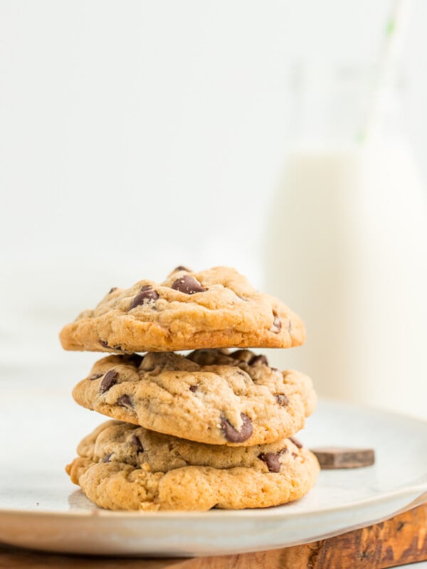 3 chocolate chip cookies stacked on a plate with a small bottle of milk with a straw in the background.