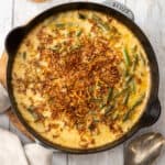 a cast iron skillet with cheesy green bean casserole topped with fried shallots on a wooden board.