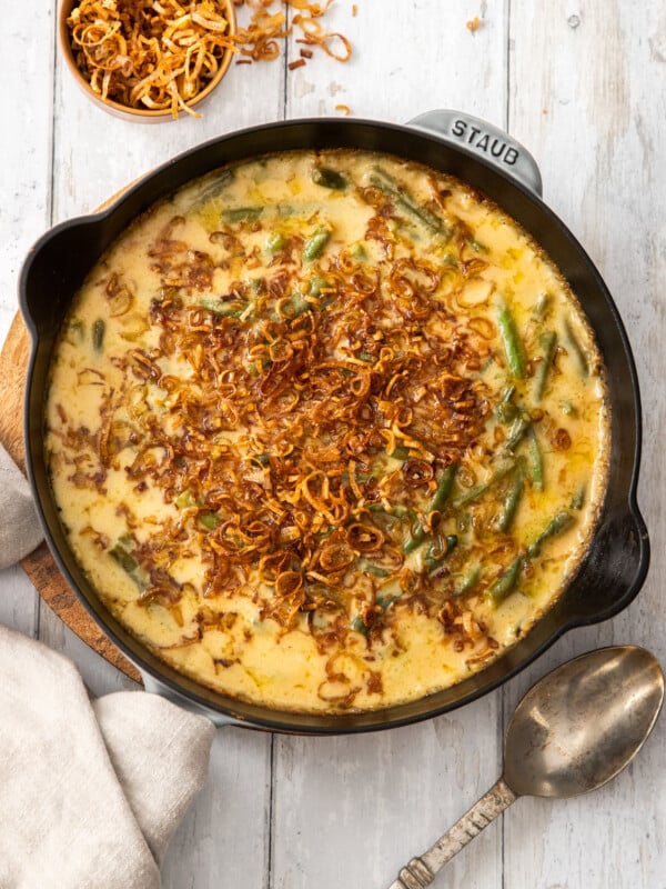 a cast iron skillet with cheesy green bean casserole topped with fried shallots on a wooden board.