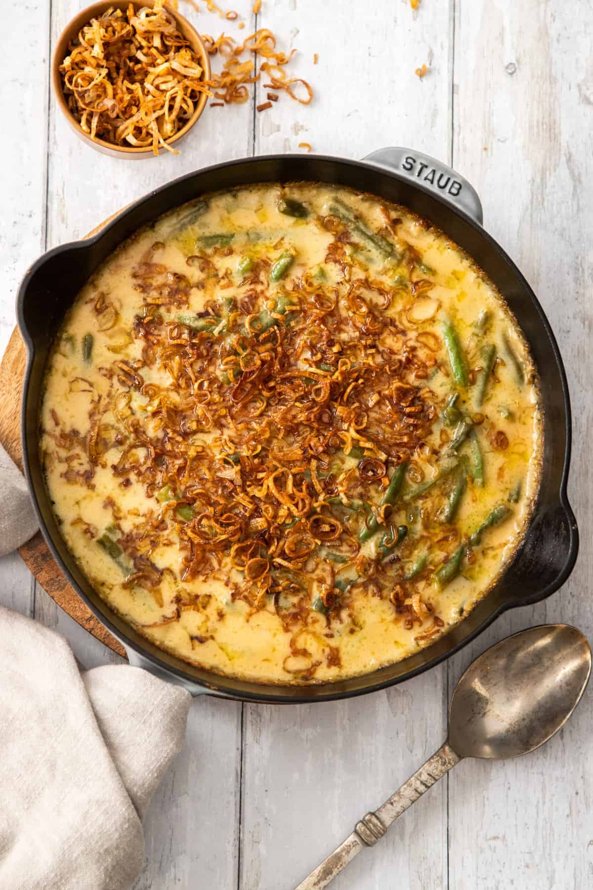 a cast iron skillet with cheesy green bean casserole topped with fried shallots on a wooden board.