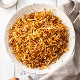 a white speckled bowl with crispy shallots surrounded by a linen, fresh shallots, and a small bowl of kosher salt.