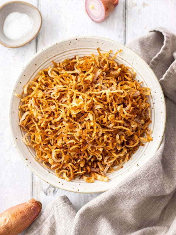a white speckled bowl with crispy shallots surrounded by a linen, fresh shallots, and a small bowl of kosher salt.