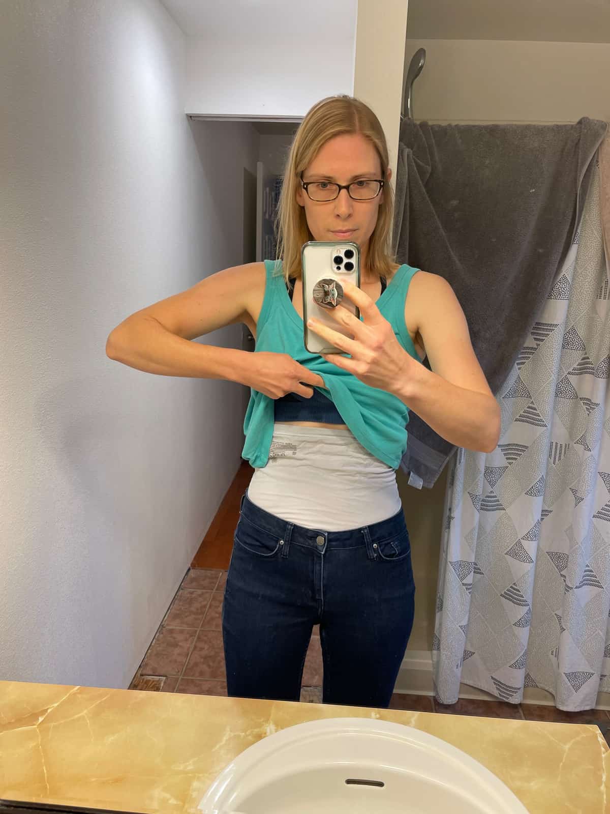 a woman in a teal tank top and glasses taking a picture in a bathroom mirror.