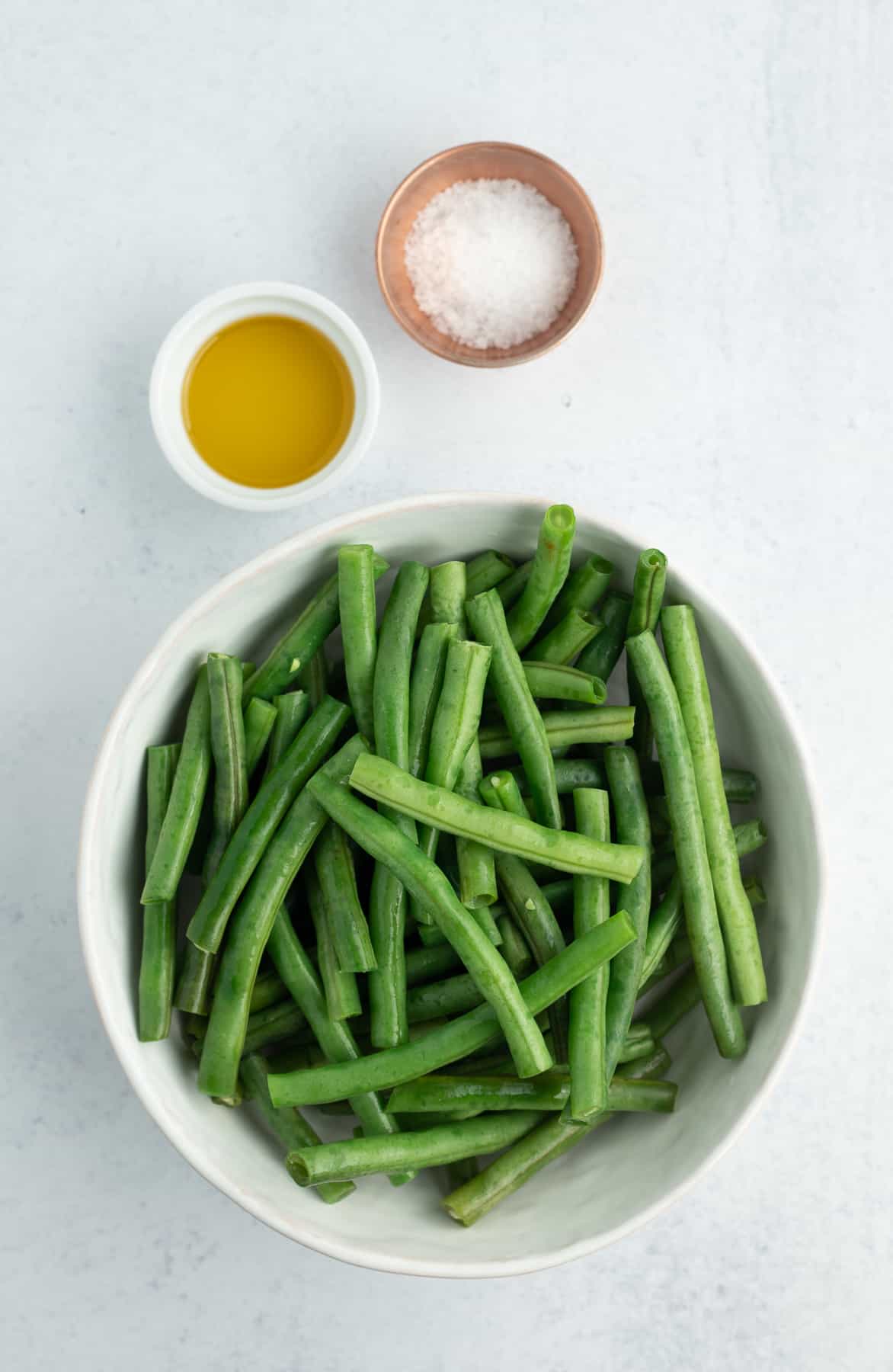 a bowl of green beans, salt, and olive oil on a white board.