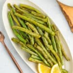 a white oval plate with roasted air fryer green beans, two lemon slices, and rose gold silverware on a white board.