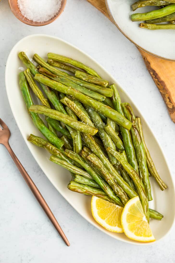 a white oval plate with roasted air fryer green beans, two lemon slices, and rose gold silverware on a white board.