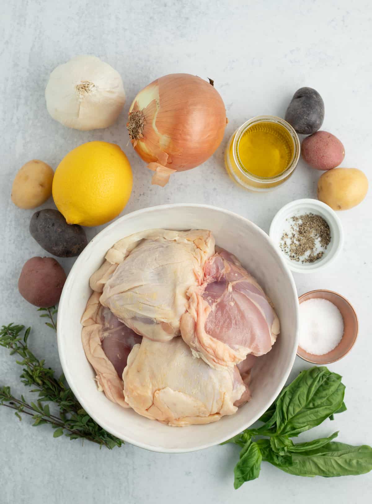 a white bowl of raw chicken thighs on a grey board with lemons, herbs, potatoes, and other ingredients.