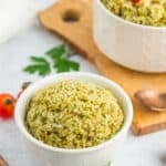 a white bowl of pesto rice with a fork, parsley, and tomatoes.