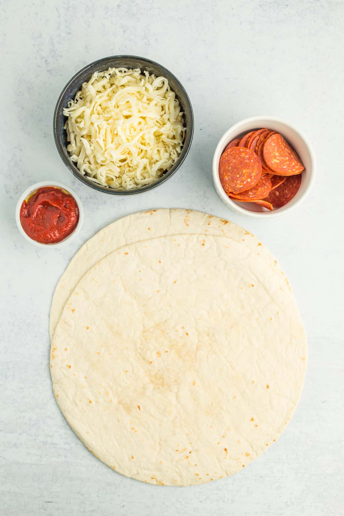 tortillas, bowls of sauce, shredded mozzarella, and sliced pepperonis.