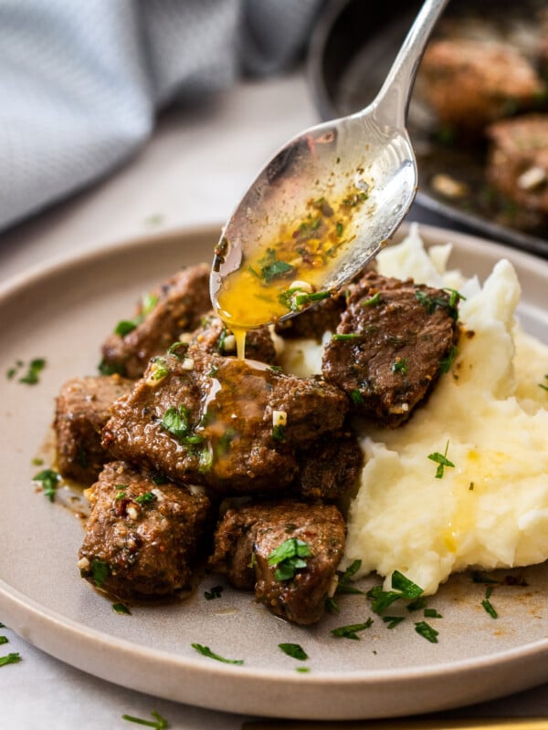 a spoonful of garlic butter being drizzled over air fryer steak bites topped with chopped parsley.