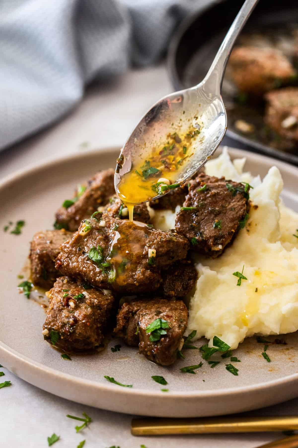 a spoonful of garlic butter being drizzled over air fryer steak bites topped with chopped parsley.
