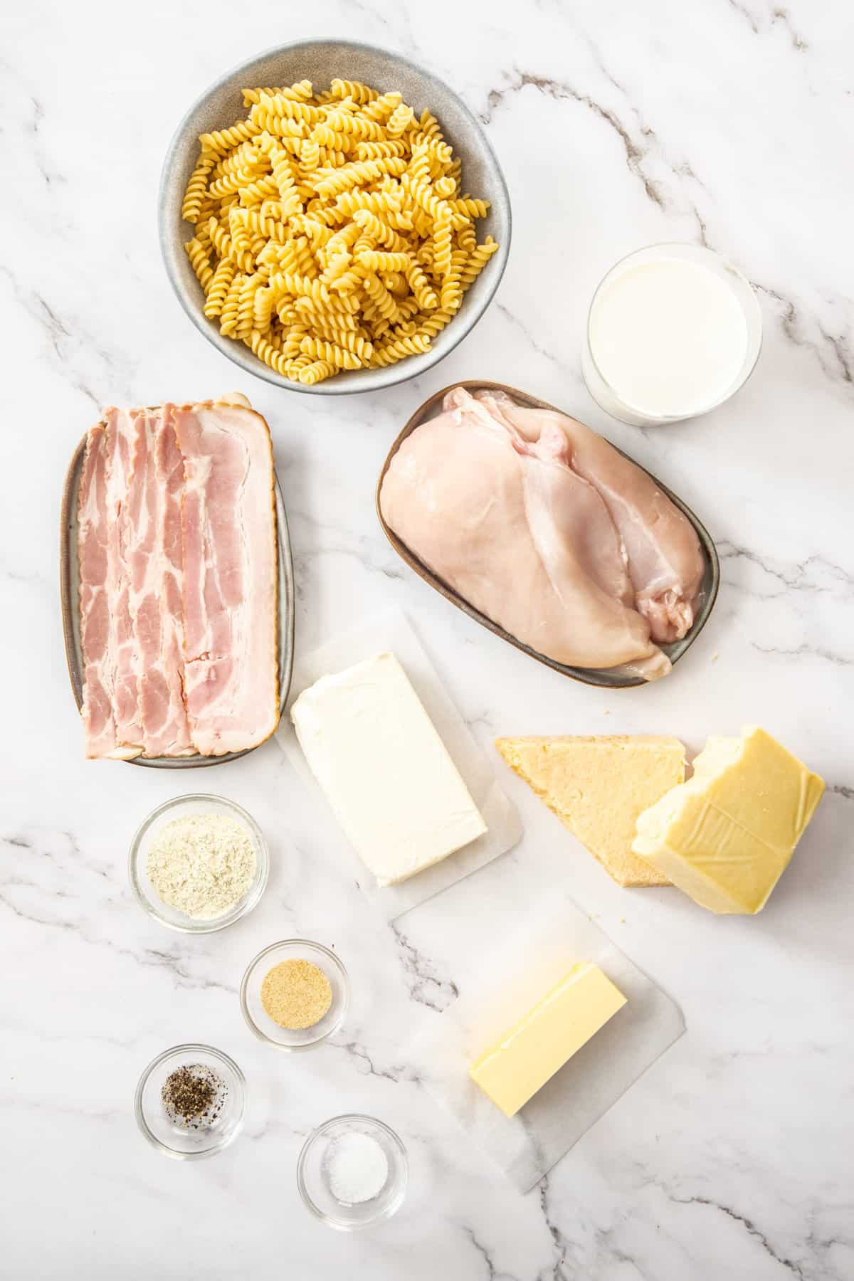 a bowl of chicken, a dish of bacon, herbs, butter, and cream cheese on a white marbled board.